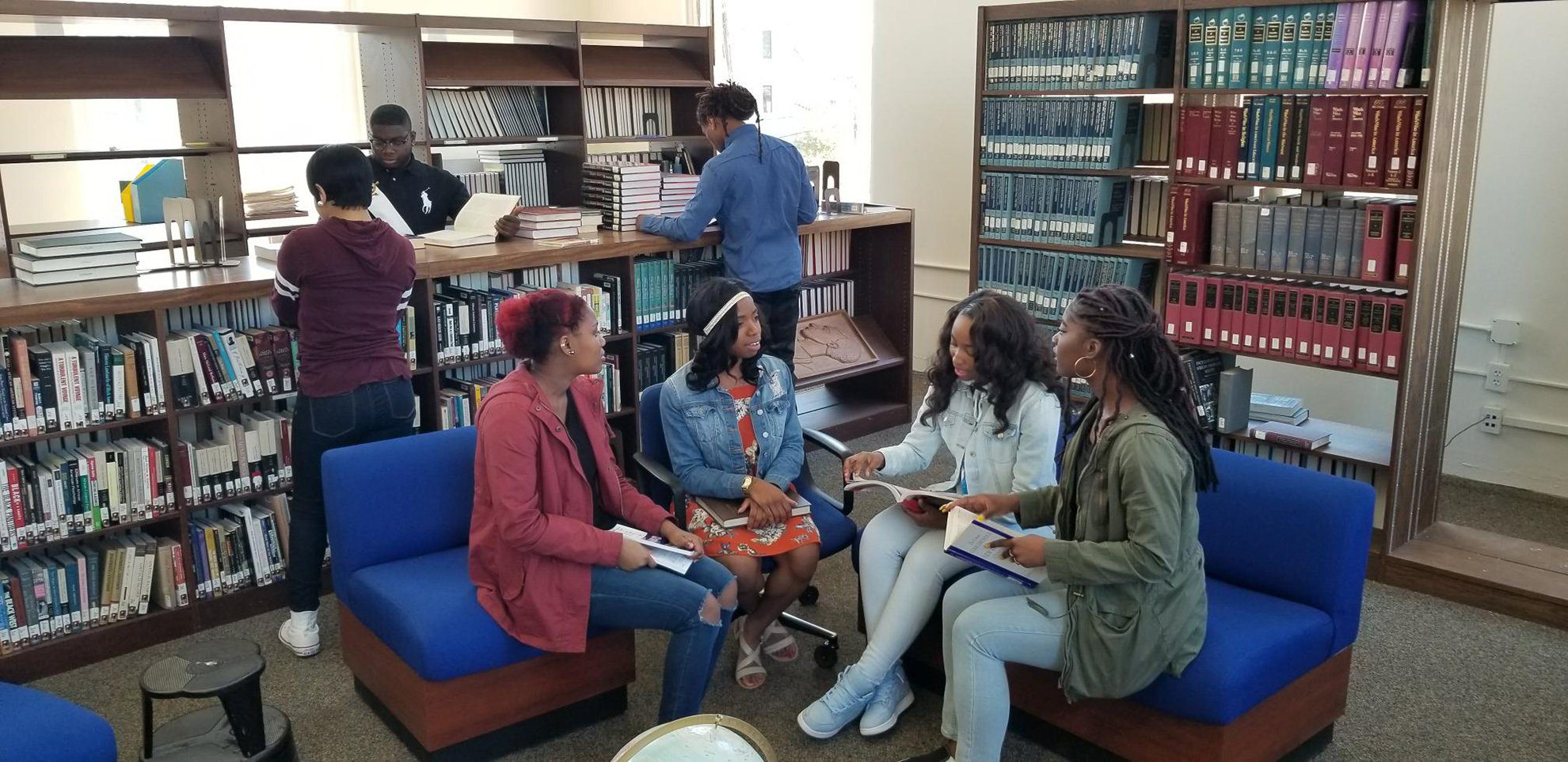 Four women sit together in a library. They are looking at books and engaged in what looks like an animated discussion. Other students can be seen checking out books in the background.