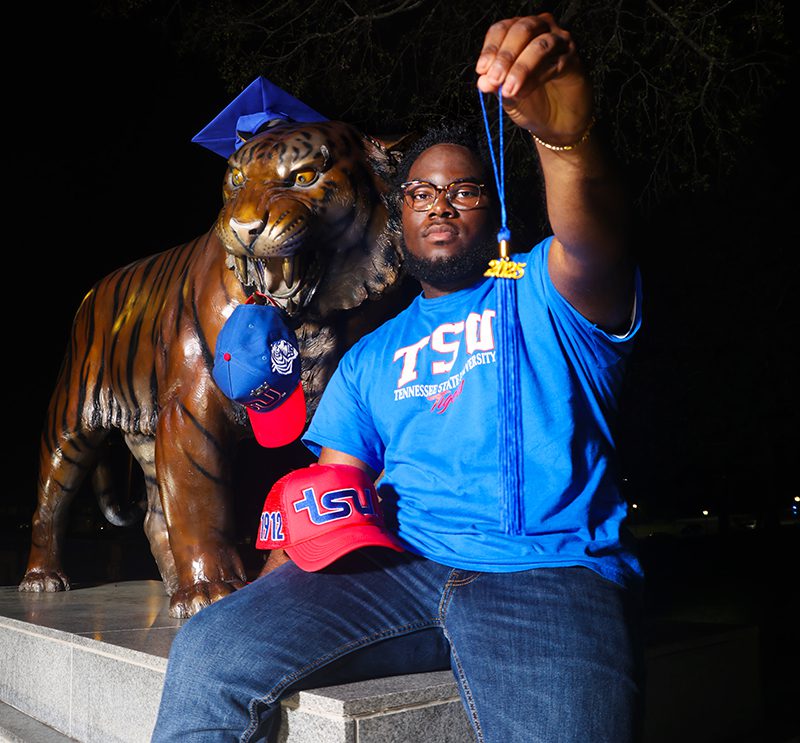 Ashton Jackson is seated next to the Tennessee State University Tiger mascot, which has a blue graduation cap on its head and a baseball cap dangles from its teeth. Ashton is wearing a blue TSU T-shirt and holding up a blue graduation cap tassel with 2025 in gold letters and a red TSU baseball hat.
