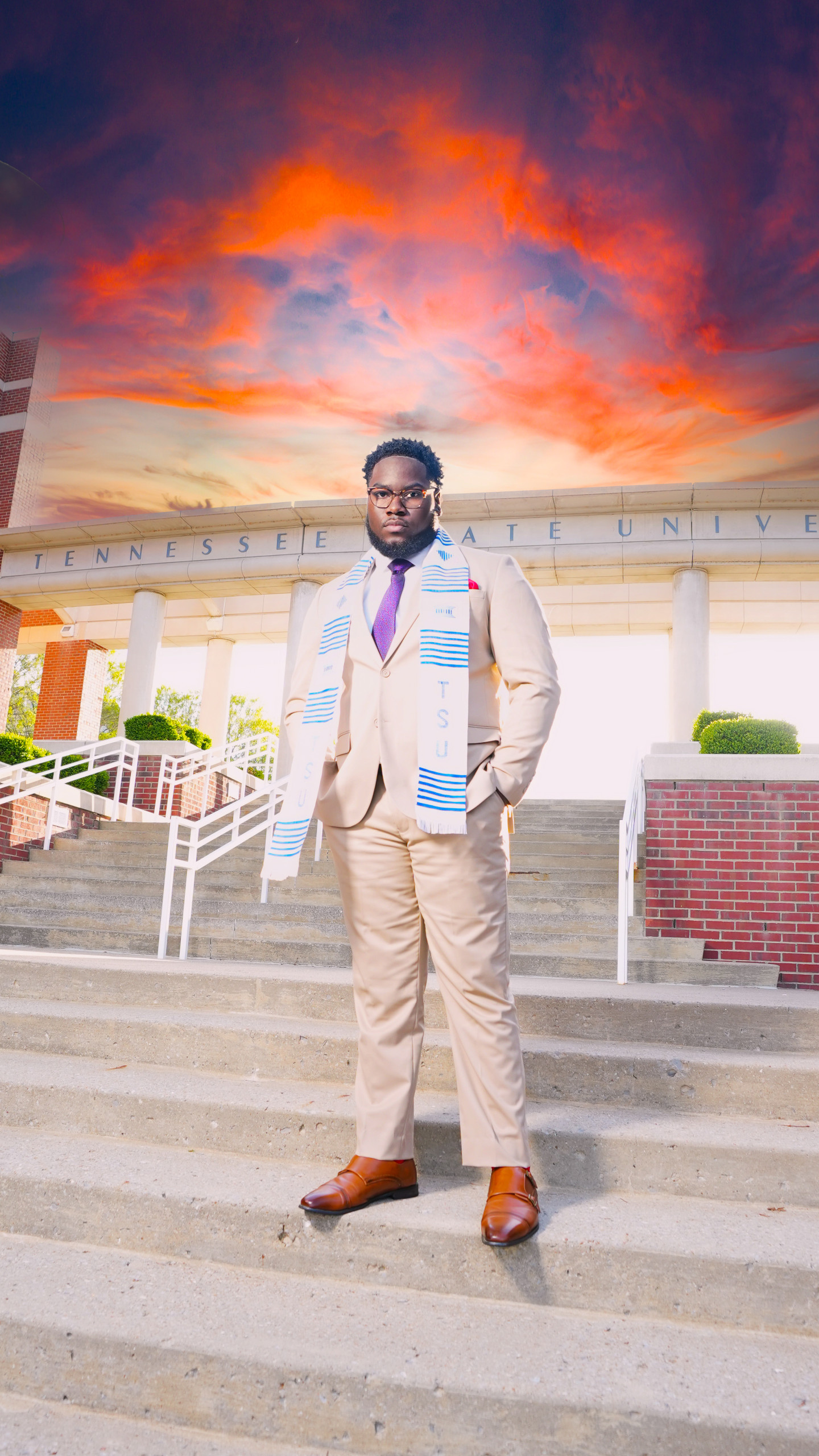 Ashton Jackson stands on stairs on campus with the words Tennessee State University engraved in a cement overhang behind him. Above him is a striking sunset with lit-up clouds in blues and oranges and purples. He is wearing a khaki suit with brown loafers, a purple tie, and a blue and white TSU scarf.