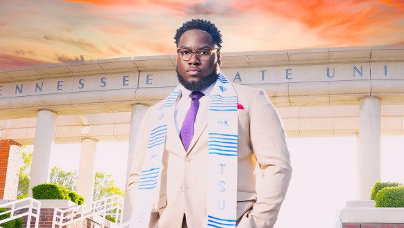 Ashton Jackson wears a khaki suit, purple tie, and a blue and white TSU scarf. Behind him, the words Tennessee State University are engraved in a cement overhang.