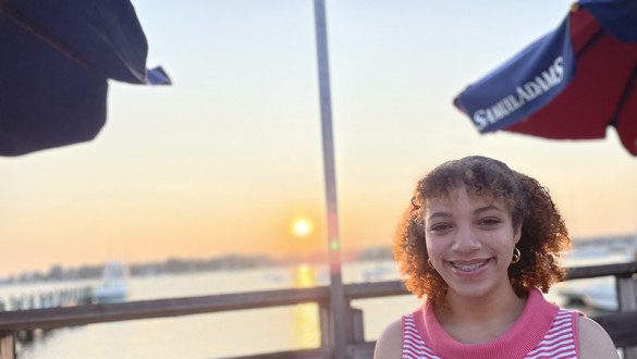 Bianca Kerr wears a pink and white striped shirt and stands in front of a sunset and body of water. The sunset reflects in the surface of the water and there are navy blue umbrellas above that say Samuel Adams.