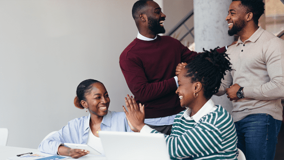 Four young african coworkers are giving high five and celebrating a successful project while working on a laptop and a tablet in a modern office