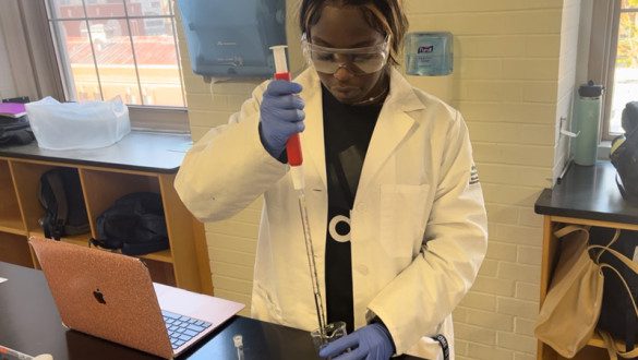 Lenah Alghali stands in a biology laboratory in a white lab coat, blue rubber gloves, and eye protection. She holds a pipette and syringe.