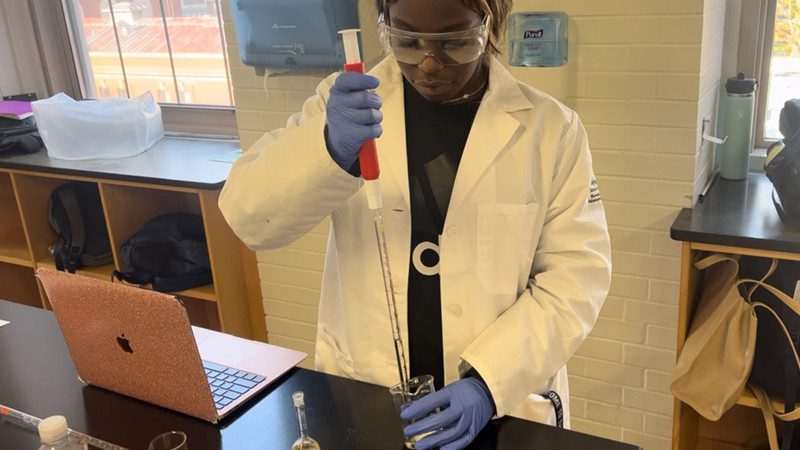 Lenah Alghali stands in a biology laboratory in a white lab coat, blue rubber gloves, and eye protection. She holds a pipette and syringe.