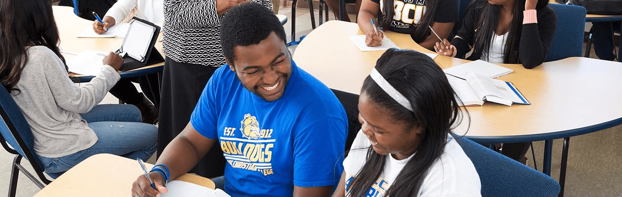 Two students sit laughing together at a desk. The desk is covered in papers and they appear to be doing homework together. Other students can be seen working at other desks in the background.