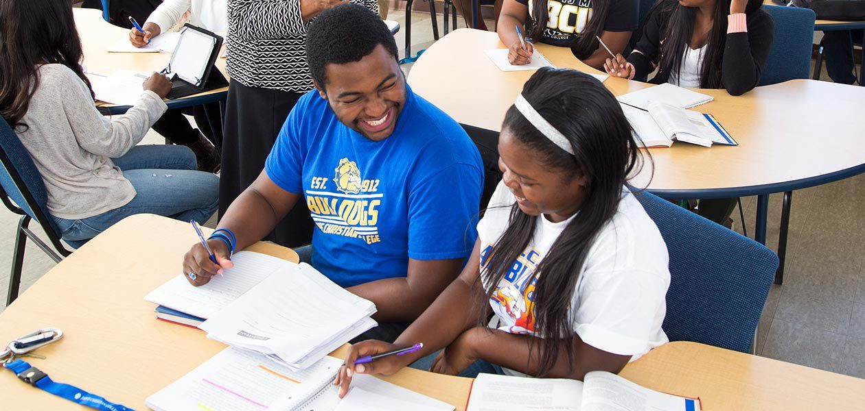 Two students sit laughing together at a desk. The desk is covered in papers and they appear to be doing homework together. Other students can be seen working at other desks in the background.