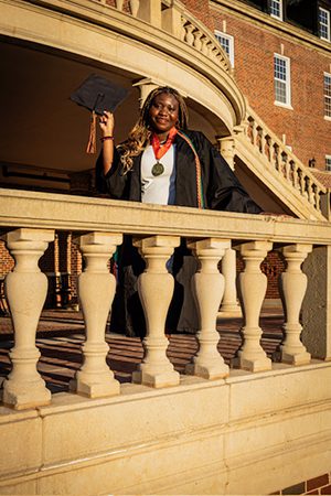 Melissa Allaro holds up her graduation cap in front of a building on campus. She is wearing her black graduation gown with a medal around her neck and colorful cords.