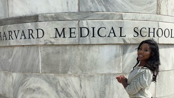 Mikaili Senwah stands in front of a granite column with the words Harvard Medical School engraved into it. She is smiling, hands clasped, wearing a black and white polka dot long-sleeved shirt.
