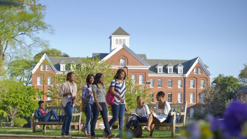 Spelman College is a historically Black college in Atlanta, GA. Students walk past a campus building in the background and greenery and flowers in the foreground.