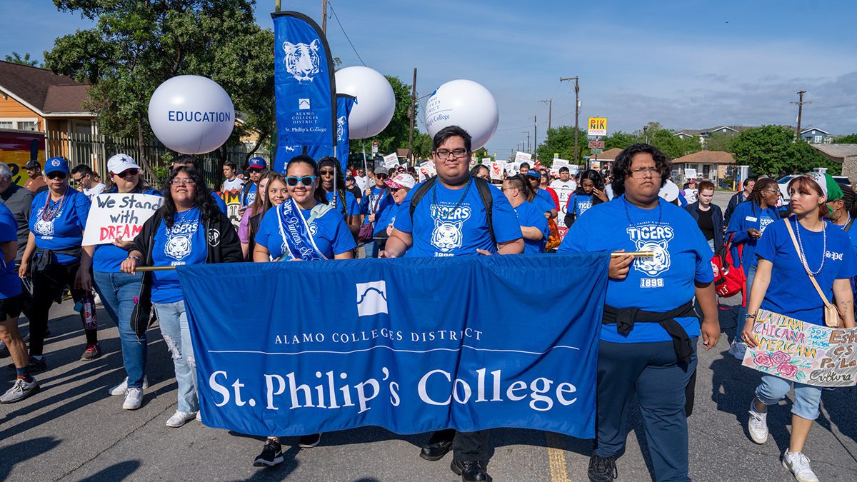 A group of students wearing matching bue t-shirts march in a parade holding a St. Philip's College banner