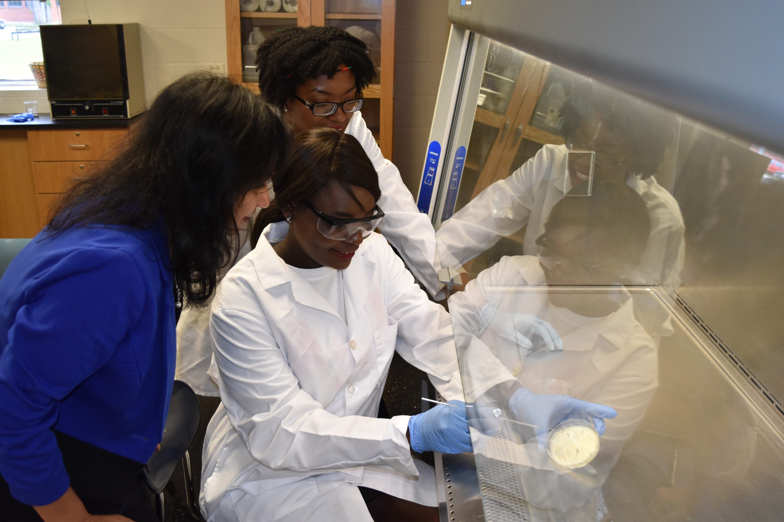 Two students in safety glasses and lab coats examine a petri dish while a woman in a blue jacket observes
