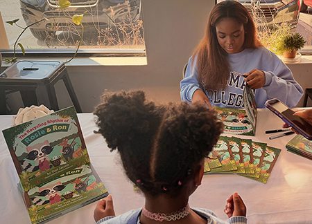 Tori Jarrett sits at a table signing her book "The Recycling Rhythm of Rosie & Ron" for a young Black girl with pigtails. Tori wears a light blue sweatshirt with Spelman College in dark blue letters on it.