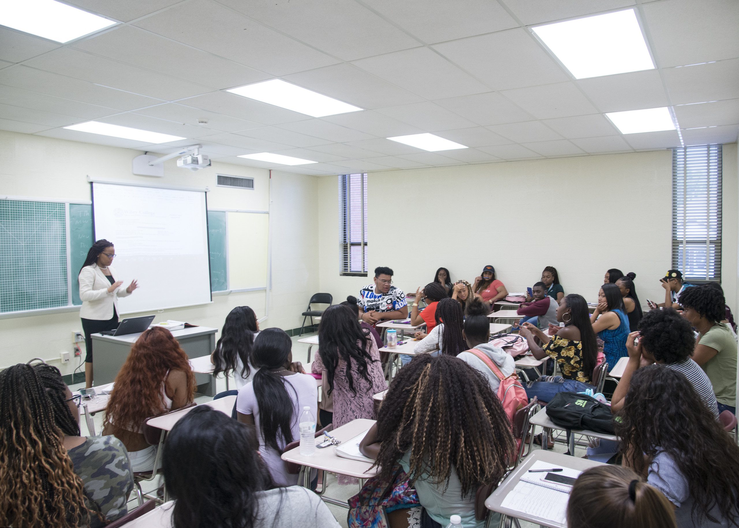 Students sit at desks in a white classroom. A teacher stands at the front of the classroom in front of a projector